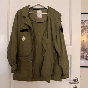 Olivgrön jacka  - Olivgrön jacka med klassisk krage, knäppning framtill och flera fickor, inklusive en med dragkedja. Jackan har patchdetaljer och en avslappnad passform, perfekt för lager-på-lager. Tillverkad i bomull för en skön känsla. Skulle säga att den passar M också. 