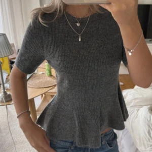 Mörkgrå stickad topp - Supersnygg mörkgrå ribbad topp med kort ärm och volang nertill. Figurnära passform som framhäver midjan och ger en feminin siluett. Perfekt till jeans eller kjol för en trendig look. Mjuk och stretchig, passar dig som gillar stilrena plagg med det lilla extra.