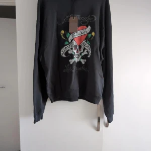 Svart Ed Hardy sweatshirt med tryck - Cool svart sweatshirt från Ed Hardy med stort färgglatt tryck på bröstet – dödskalle, hjärta och texten 'Love Kills Slowly'. Tröjan har långa ärmar och ribbade muddar. Perfekt för dig som gillar streetstyle och statement-plagg.