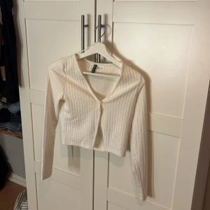 Vit ribbad croppad topp H&M Divided - Säljer en vit croppad topp från H&M Divided i storlek S. Toppen är ribbad med lång ärm och har en enkel knäppning framtill. Perfekt att styla med jeans eller kjol för en clean look.