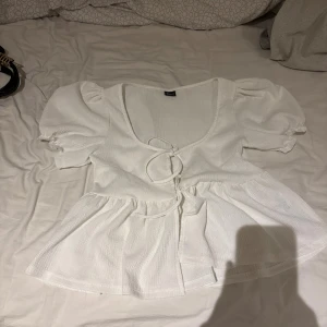 Vit blus med puffärm från Gina Tricot - Säljer en vit blus från Gina Tricot i storlek S. Blusen har puffiga korta ärmar, v-ringad hals och två knytband framtill. Materialet är lätt strukturerat och ger en luftig känsla. Perfekt för dig som gillar romantiska detaljer och volangkant. Bara provat 1 gång, som ny. Pris kan diskuteras 