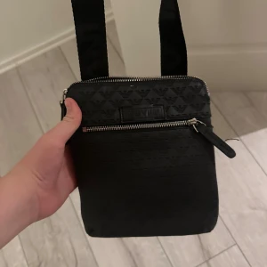  Armani Väska - Snygg svart axelväska från Emporio Armani med justerbart axelband och silverfärgade dragkedjor. Väskan har diskret mönster med loggor och en praktisk ytterficka framtill. Perfekt storlek för det viktigaste och stilren design med tydlig Armani-logga.