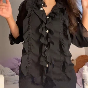 Svart volangskjorta med knappar - Svart söt skjorta med volanger längs knäppningen och spetsdetaljer på framsidan. Skjortan har pärlemorknappar och är tillverkad i ett lätt, transparent tyg med trekvartsärm. Perfekt för dig som gillar en extra touch på outfiten.