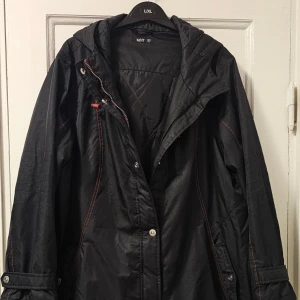 Svart vindjacka med röda detaljer - Svart vindjacka från VIT i storlek L/XL. Jackan har röda kontrastsömmar, huva med dragsko och praktiska fickor med knappdetaljer. Stängs med både dragkedja och tryckknappar. Perfekt för blåsiga dagar och enkel att styla till vardags.