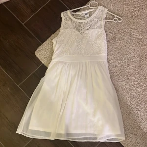 Vit spetsklänning från Vero Moda - Superfin vit kort student klänning från Vero Moda med spetsdetaljer upptill och vackert fall i kjolen. Klänningen är ärmlös och har en djup v-ringning i ryggen med transparent spets. St M.