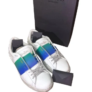 Valentino Garavani Open  - Snygga Valentino Garavani Open sneakers i vitt skinn med en bred, glansig panel i blått och grönt över mitten. Klassisk rund tå, platt sula och grå snörning. Ikoniska nitar baktill och logga på hälen. Inget finns med tyvärr då de försvunnit men skorna är självklart äkta!! Rätt sunkiga därav pris