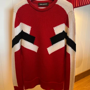 Röd stickad tröja Neil Barrett - Cool röd stickad tröja från Neil Barrett med grafiskt mönster i svart och vitt. Tröjan har rund halsringning, långa ärmar och en slim fit passform. Perfekt för dig som vill sticka ut med en unik design och snygga kontraster.