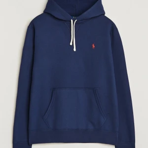 Marinblå hoodie från Polo Ralph Lauren - Snygg marinblå hoodie från Polo Ralph Lauren med klassisk huva och vit snörning. Tröjan har en stor magficka och den ikoniska röda Polo-loggan broderad på bröstet. Perfekt för chill dagar och enkel att matcha med jeans eller joggers. M passar S, saknar snörena