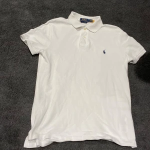 Vit pikétröja från Polo Ralph Lauren - Klassisk vit pikétröja från Polo Ralph Lauren med korta ärmar och blå broderad logga på bröstet. Tröjan har krage och knäppning med två knappar. Perfekt för en clean och stilren look.