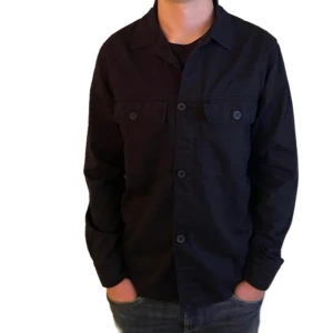 OANVÄND overshirt med knappar mörkblå M - Stilren mörkblå overshirt med klassisk krage och knäppning framtill som är helt OANVÄND. Två bröstfickor med lock och knappar