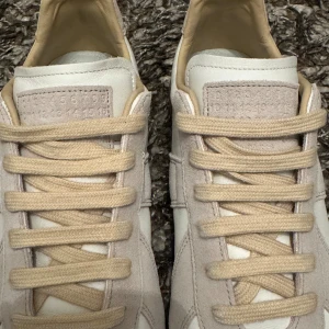 Maison Margiela sneakers beige/vit - Maison Margiela sneakers i beige och vitt med klassisk gummisula. Ovandel i skinn och mocka, beige snörning och diskret logotyp på plösen och sulan. Snygg minimalistisk design som passar till allt.