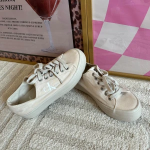 Beige sneakers från Calvin Klein Jeans - Snygga beige sneakers från Calvin Klein Jeans med vit logga på sidan. Skorna har rund tå, platt sula och snörning. Tillverkade i canvasmaterial som ger en clean och fräsch look. Perfekta för dig som gillar stilrena och klassiska sneakers.