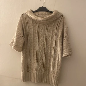 Beige stickad polotröja  - Säljer en beige stickad polotröja i storlek M. Tröjan har korta ärmar, bred polokrage och snygga kabelstickade detaljer framtill. Den är ribbad vid ärmslut och nederkant, perfekt för dig som gillar mysiga och trendiga plagg.