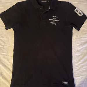 Svart pikétröja Peak Performance - Svart pikétröja från Peak Performance med vit logga på bröstet och en stor siffra 86 på ärmen. Klassisk krage, korta ärmar och knäppning framtill. Perfekt för en clean och sportig look.