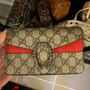  axelväska med GG-mönster - Säljer en  axelväska i beige och brunt GG-mönster med röda mockadetaljer och silverfärgat spänne. Väskan har en kedjerem och är tillverkad i canvas och mocka. Perfekt för dig som vill ha en ikonisk och trendig väska med lyxig känsla.