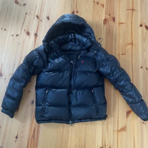 Svart pufferjacka från Polo Ralph Lauren - Snygg svart pufferjacka från Polo Ralph Lauren med huva och dragkedja. Jackan har två fickor med dragkedja och klassisk röd broderad logga på bröstet. Perfekt för kalla dagar och riktigt skön att bära. Materialet är glansigt och känns lyxigt. Köpt förra året för 6695kr men har inget kvitto kvar, jackan är knappt använd då jag inte gillade helt hur den satt och är även sugen på en mörkgrön moncler maya, kan vara öppen för byten och pris kan diskuteras vid snabb affär🤩