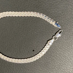 Silver pläterade armband i kedjemodell 925 - Stilrent armband i silverfärg med platt, bred kedja och snyggt mönstrad yta. Armbandet har en klassisk karbinhake och är tillverkat i pläterad silver, vilket syns på stämpeln 925. Perfekt för dig som gillar en clean och tidlös look. längd, 21 bredd, 5mm