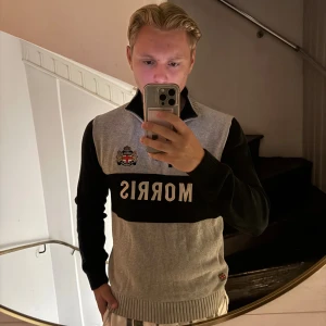 Grå och svart half zip tröja Morris - Säljer en snygg grå och svart stickad tröja från Morris med half zip och broderad logga på bröstet. Tröjan har ribbad krage, muddar och en bred svart panel med texten MORRIS framtill. Perfekt för dig som gillar klassisk stil.