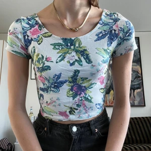 Blommig croppad topp med kort ärm - Superfin croppad topp med blommigt mönster i blått, grönt, rosa och lila på vit botten. Toppen har rundad halsringning både fram och bak samt korta ärmar. Perfekt för dig som gillar färg och mönster! Materialet känns mjukt och stretchigt.