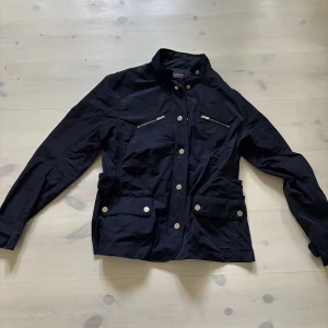 Fieldjacket Från Snoot - Snygg svart fieldjacket från Snoot med ståkrage, silverfärgade knappar och dragkedjor på bröstet. Jackan har två stora fickor med lock och knapp framtill, samt broderad logga på insidan. Perfekt för dig som gillar stilren och funktionell design. Det är L men sitter som M