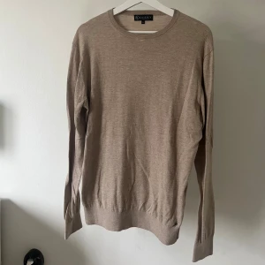 Beige stickad tröja från Therlunds - En stilren beige stickad tröja från therlunds egna märke ”LAGENCY” i klassisk modell med rund halsringning och långa ärmar. Tröjan har ribbade muddar vid ärmslut och nederkant, vilket ger en snygg passform. Perfekt för lager-på-lager under kyliga dagar.