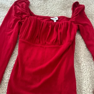 Röd ribbad blus från Bubbleroom - Säljer en snygg röd blus från Bubbleroom i storlek XS. Blusen har ribbad struktur, långärmade puffärmar och en smickrande fyrkantig ringning med rynkade detaljer upptill. Tar frakt, använd en gång 