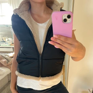 Svart dunväst med beige foder - Snygg svart dunväst med mjukt beige teddyfoder och dragkedja framtill. Västen har en hög krage och quiltad design som ger en cool look. Perfekt att slänga över en t-shirt för extra värme och stil. Passar dig som gillar att hålla det enkelt men ändå trendigt.