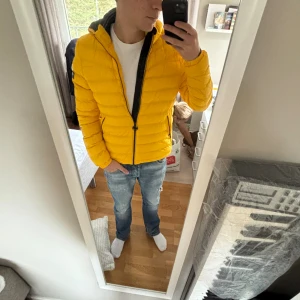 Gul pufferjacka med huva - Säljer en riktigt snygg gul pufferjacka med huva och svarta detaljer. Jackan har dragkedja framtill och två fickor med dragkedja på sidorna. Perfekt för dig som vill sticka ut med en färgglad och modern look.