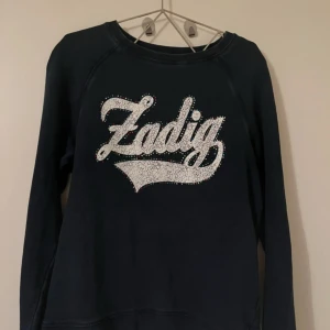 Marinblå sweatshirt från Zadig & Voltaire - Cool marinblå sweatshirt från Zadig & Voltaire med stort, vitt tryck och färgglada små nitar runt texten på bröstet. Tröjan har rund hals och långa ärmar, perfekt för en avslappnad och trendig look. Är i barnstorlek 16 men sitter som en S