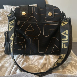 Svart axelväska från FILA med mönster - Cool svart axelväska från FILA med grafiskt mönster i guld och bred axelrem med logga. Väskan har både handtag och justerbar rem, dragkedjor på sidorna och detaljer i guld. Perfekt för dig som vill ha en sportig men trendig look. Ny kostar den 400 men säljer för 200 