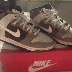 Nike Dunk High i beige och brunt mocka - Nike Dunk High sneakers i en snygg mix av beige och brunt mocka med vita detaljer och snörning. Skorna har en klassisk hög siluett och vit swoosh på sidan. Perfekta för dig som gillar streetwear och vill ha något stilrent men ändå unikt.