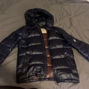Mörkblå Moncler dunjacka med huva - Snygg mörkblå dunjacka från Moncler med huva och bruna detaljer på insidan. Jackan har två dragkedjeförsedda fickor, quiltad design och coolt serietryck på fodret. Perfekt för kalla dagar och har en modern look som sticker ut. Kontakta vid frågor.