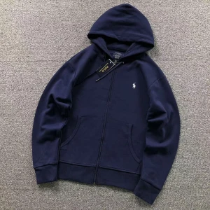 Marinblå hoodie från Polo Ralph Lauren - Snygg marinblå hoodie från Polo Ralph Lauren med huva och dragkedja. Tröjan har två fickor framtill och den klassiska vita Polo-loggan broderad på bröstet. Perfekt för chill dagar och enkel att matcha med jeans eller joggers.