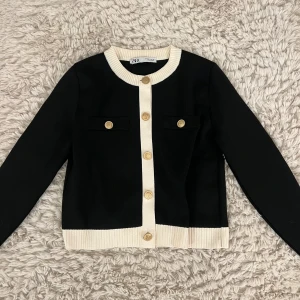 Svart kofta med guldknappar - Säljer en svart kofta från Zara med vita ribbade kanter och snygga guldiga knappar. Koftan visar inget spår av användning. 