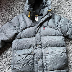 Grå dunjacka från Fjällräven med huva - Fjällräven storlek xl  På armen och längst ner på ryggen finns de två lagningar se bilderna i övrigt är jackan i gott skick 