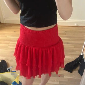 Röd volangkjol från Lindex - Superfin röd kortkjol från Lindex med bred smock i midjan och flera lager volanger. Kjolen är gjord i återvunnen polyesterblandning och har en luftig, plisserad känsla som ger extra rörelse. Perfekt för dig som vill sticka ut med färg och volym.