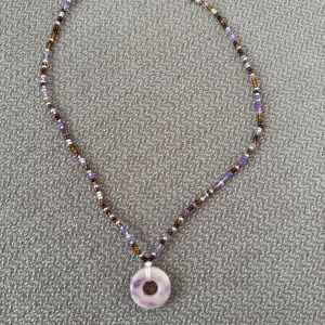 Pärlhalsband med lila cirkel-hänge - Unikt halsband med små pärlor i lila, brunt, vitt och gult. Halsbandet har ett cirkelformat hänge i lila och vitt, den är 44cm långt 💜🤍🤎