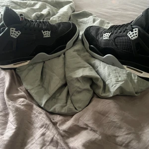 Svarta Nike Air Jordan 4 sneakers - Säljer ett par svarta Nike Air Jordan 4 sneakers med grå och vita detaljer samt röd logga under sulan. Skorna har meshpaneler, snörning och den klassiska Jumpman-loggan på hälen. Ovandelen är i mocka och textil, och sulan är i gummi för grym komfort. Knappast använda och perfekta inför vintern 