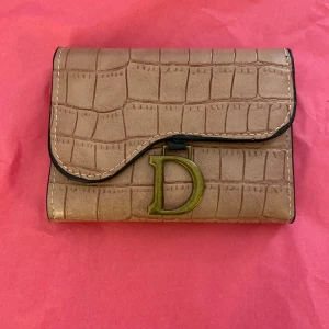 Rosa plånbok med krokopräglat mönster - Snygg beige plånbok i krokopräglat skinnimitation med stor guldfärgad D-detalj framtill. Plånboken har flera fack för kort och ID samt tryckknappsstängning. Perfekt för dig som vill ha en stilren och trendig accessoar i väskan.