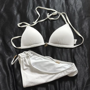 Vit ribbad bikini med snörning - Superfin vit bikini med ribbad struktur och snygga snörningsdetaljer. Toppen har trekantsformade kupor och ett litet hjärtformat smycke framtill. Underdelen har justerbara band i sidorna för perfekt passform. Perfekt för stranden eller poolen!