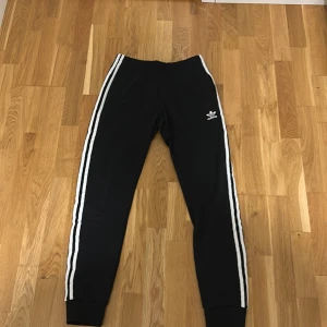 Svarta Adidas byxor med vita ränder - Svarta Adidas byxor med klassiska tre vita ränder längs benen och broderad logga på höften. Modellen har resår i midjan och muddar vid bensluten. Tillverkade i återvunnet Primeblue-polyestermaterial. Perfekta för en sportig och avslappnad look.