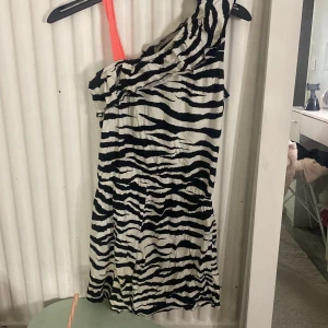 Zebramönstrad one shoulder klänning - Snygg kort klänning med zebramönster i svart och vitt. Klänningen har en trendig one shoulder-design med en axelbandslös sida och ett orange axelband. Perfekt för dig som vill sticka ut med en cool och djärv look.