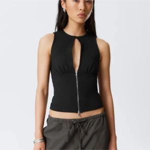 Svart ärmlös topp med dragkedja - Snygg svart ärmlös topp med rund hals och djup öppning framtill som stängs med en liten knapp upptill. Dragkedja går från bröstet till nederkant och ger en cool vibe. Croppad passform som visar midjan, perfekt för att styla med höga byxor.