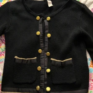 Svart kofta med guldknappar från Vila - Svart stickad kofta från Vila med snygga guldknappar framtill och två fickor med dekorativ kant. Koftan har rund hals och detaljer i svart tyg längs knäppningen och fickorna. Perfekt för dig som gillar klassisk stil med en twist.