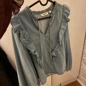 Blå volangblus från Kappahl - Supersöt ljusblå blus från Kappahl med volanger längs axlar och bröst, v-ringning med knytband och långa ärmar. 