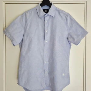Blå kortärmad skjorta från G-Star Raw - Ljusblå, kortärmad skjorta från G-Star Raw i storlek M. Klassisk krage och vita knappar framtill. Bomull med ett diskret mönster i vitt.