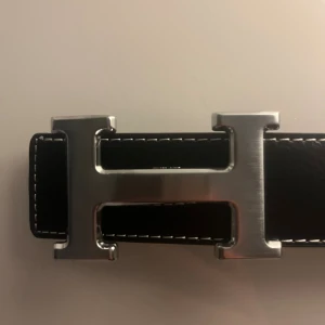 Svart H-bälte - Svart bälte i skinn med ikoniskt H-format metallspänne i silver. Bältet har vita kontrastsömmar längs kanterna och en klassisk, stilren design som passar till jeans eller kostymbyxor. Skriv om du har frågor!😁TAR EMOT BYTEN