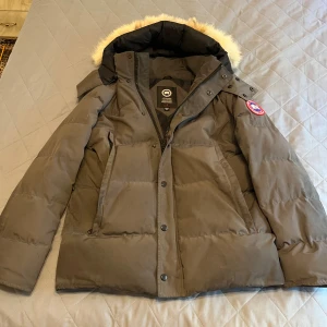 Grå Canada Goose Wyndham dunjacka med päls - Säljer en grå dunjacka från Canada Goose med ikonisk patch på ärmen och fluffig päls runt huvan. Jackan har dragkedja, knappar och två stora fickor framtill. Perfekt för kalla dagar och har en clean, klassisk look.