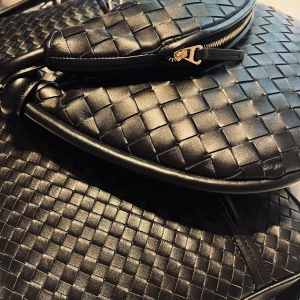 Svart flätad axelväska i skinn - Snygg svart axelväska i flätat skinn med rundad form och bred axelrem. Väskan har en dragkedja framtill och coola knutdetaljer på remmen. Perfekt för dig som gillar stilrena accessoarer med lite edge.