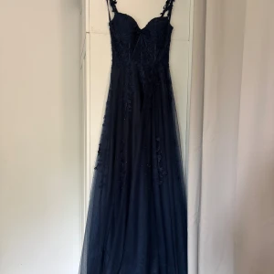 Marinblå långklänning med spetsdetaljer - Säljer en marinblå långklänning med tunna axelband och v-ringad urringning. Klänningen har vackra spetsdetaljer och broderade blommor på livet samt ett skirt lager i tyll som ger en drömmig känsla. Perfekt för bal eller student. Sitter snyggt och har en elegant siluett.